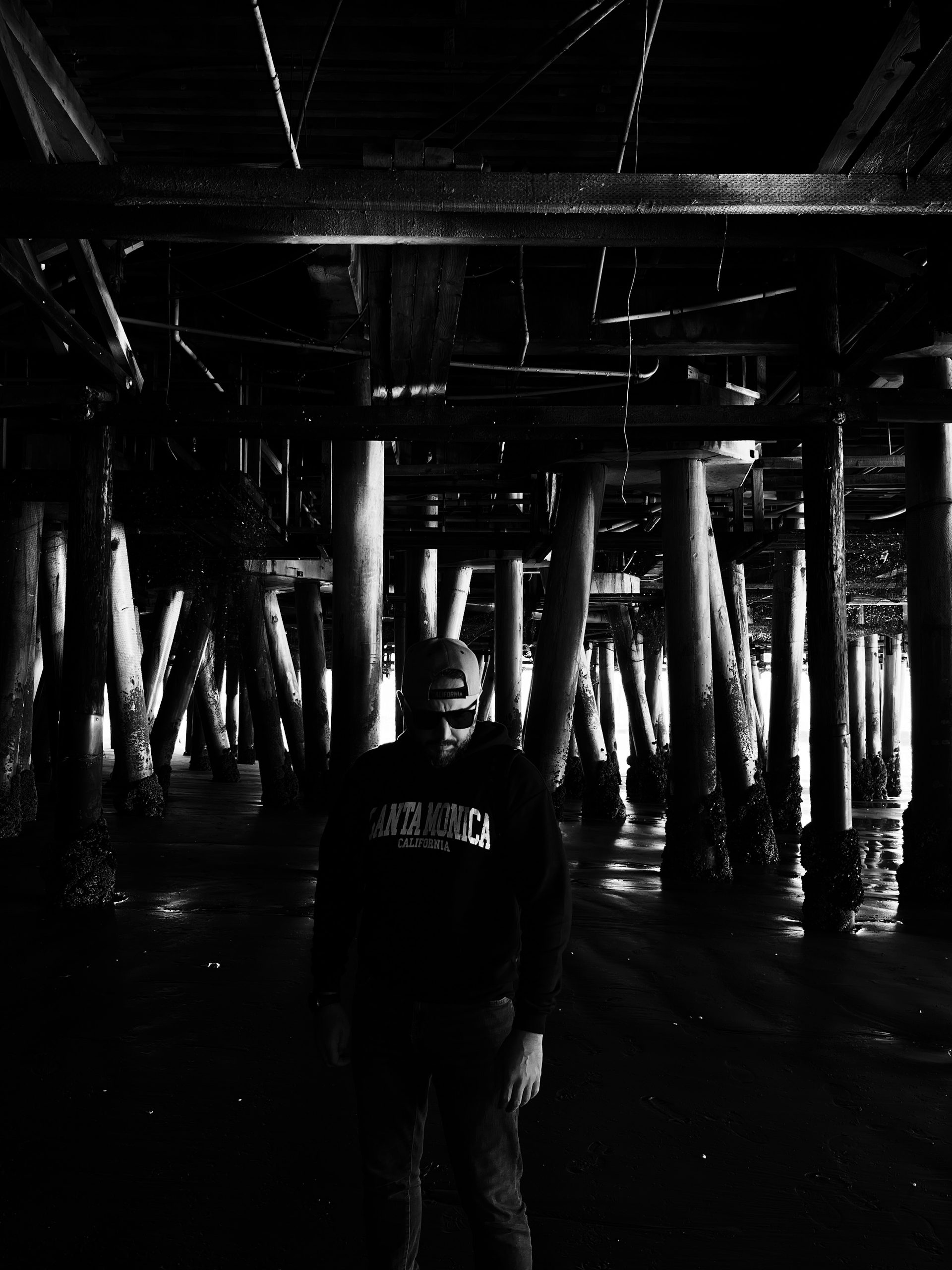 Muzaffer Keskin standing beneath the Santa Monica Pier, high-contrast black and white